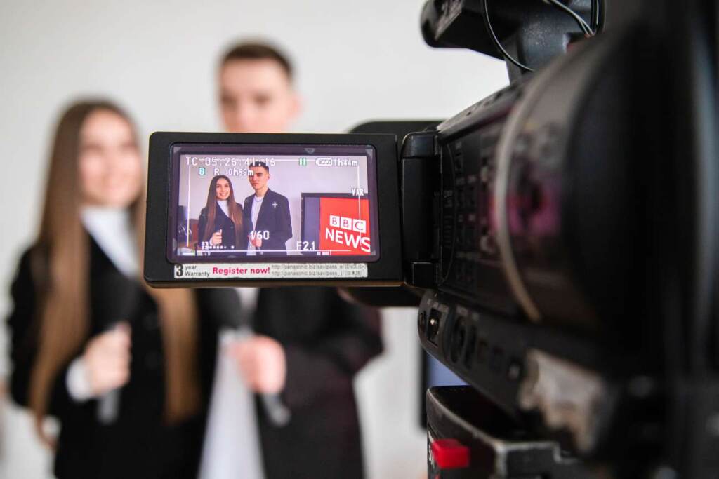 на передньому плані є камера, а на екрані камери стоять дівчина і чоловік, ймовірні журналісти, біля них є екран де є напис "BBS news"